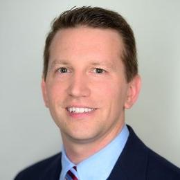 Man with dark hair wearing suit light dress shirt and tie in front of beige wall