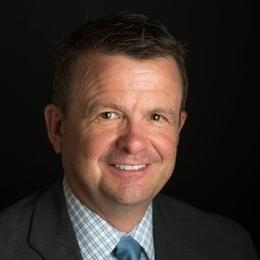 Man with dark hair wearing dark suit with plaid dress shirt and blue tie