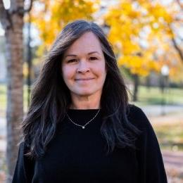 Lady with long dark hair wearing black sweater and necklace standing in front of yellow fall tree on campus